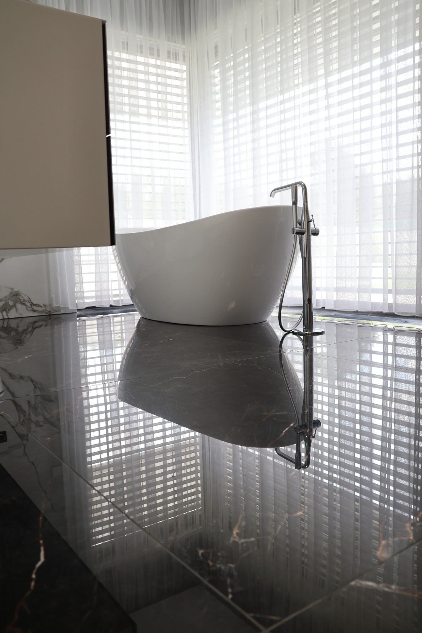 Nowoczesna, wolnostojąca wanna z chromowaną baterią na tle okna z białymi, przezroczystymi zasłonami i błyszczącej, ciemnej podłogi z marmurkowym wzorem.