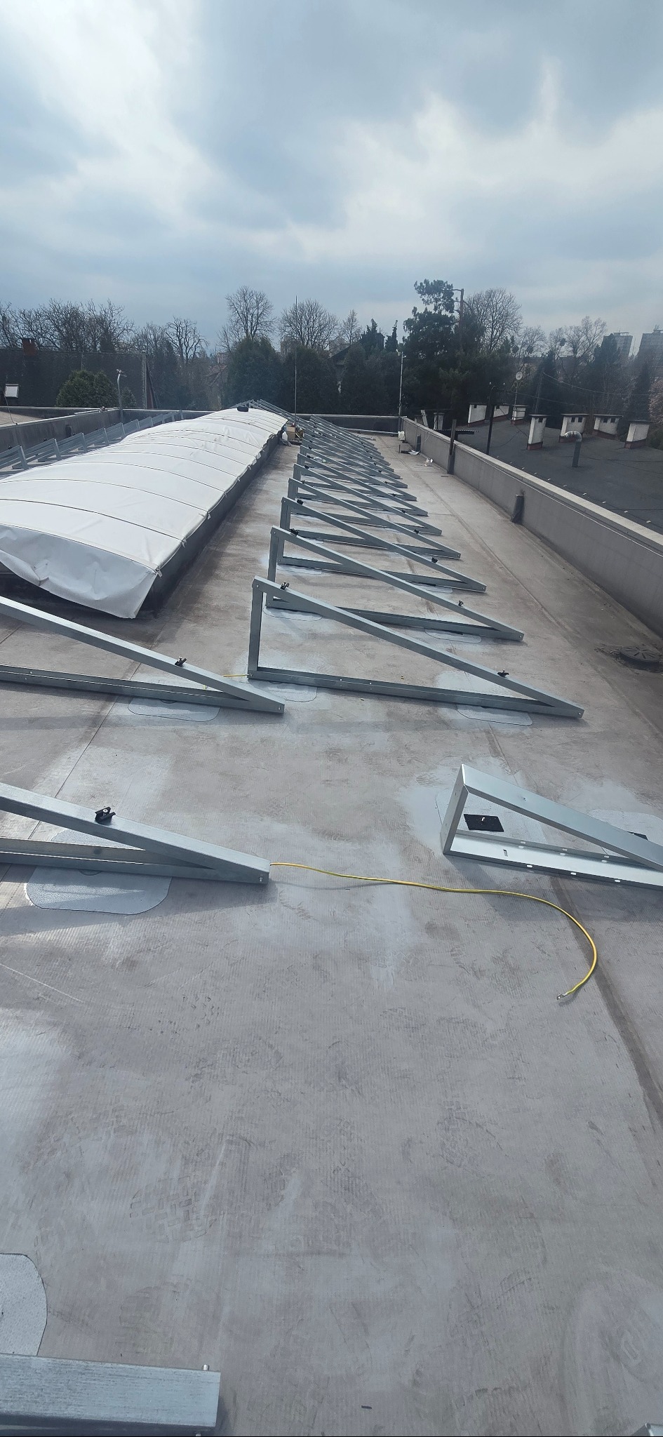 Montaż stelażu pod panele słoneczne na dachu, widoczne metalowe konstrukcje i przewody. Częściowo zainstalowana osłona. Pochmurne niebo w tle.