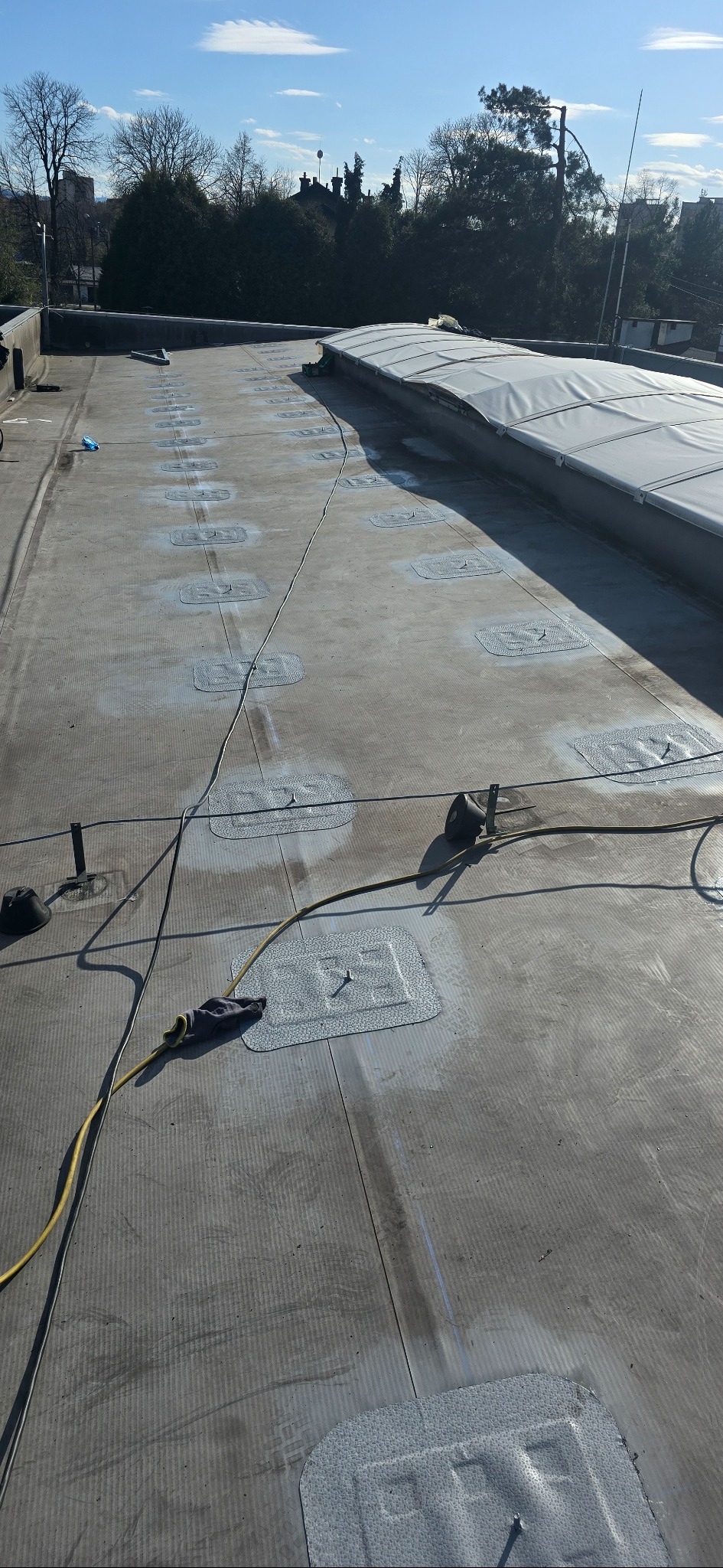 Montaż instalacji na dachu: widoczne uchwyty mocujące, kable i fragment instalacji. Dach pokryty papą, w tle drzewa i błękitne niebo.