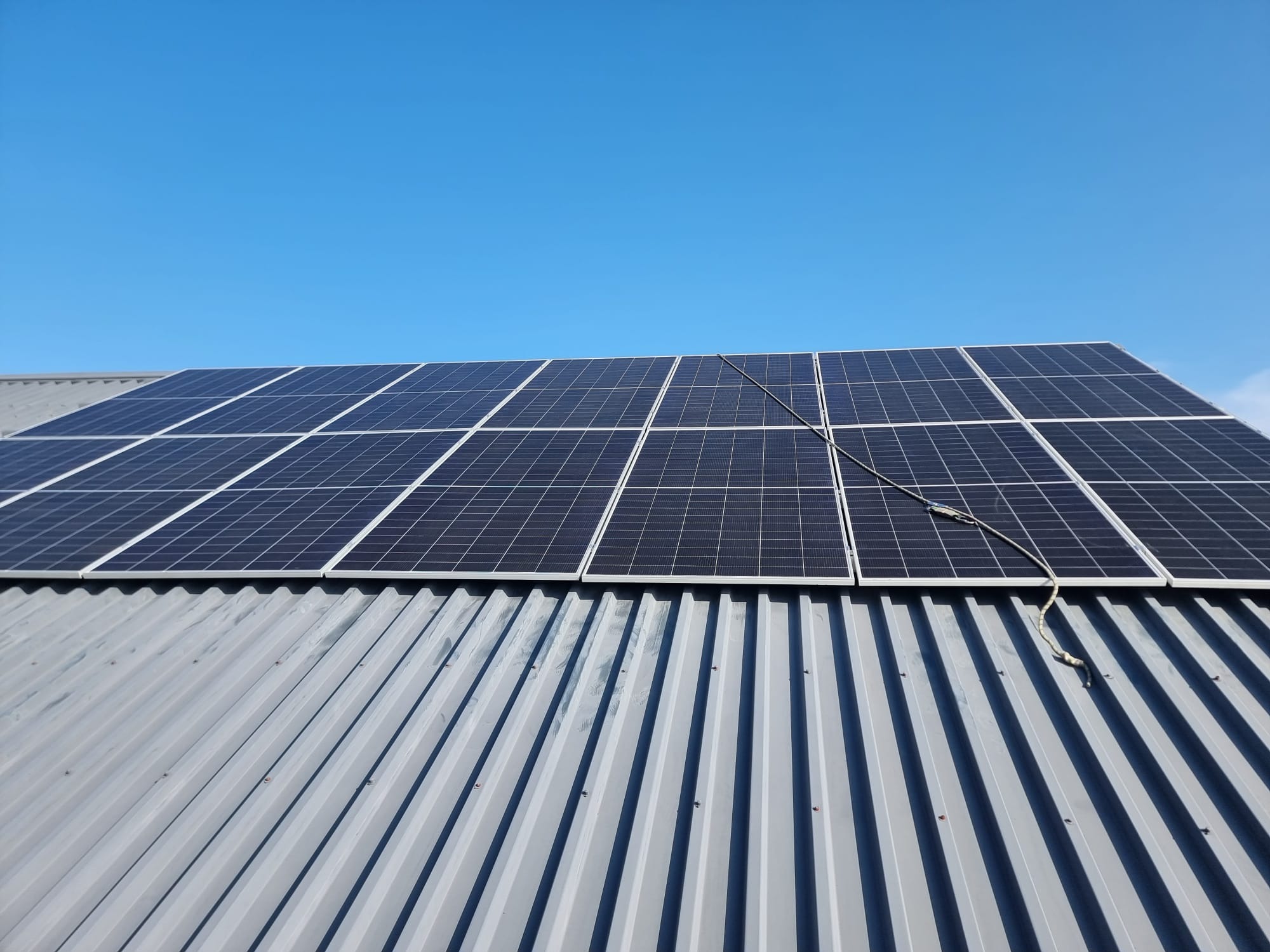 Panele słoneczne na blaszanym dachu budynku w słoneczny dzień. Widoczne kable. Instalacja fotowoltaiczna.