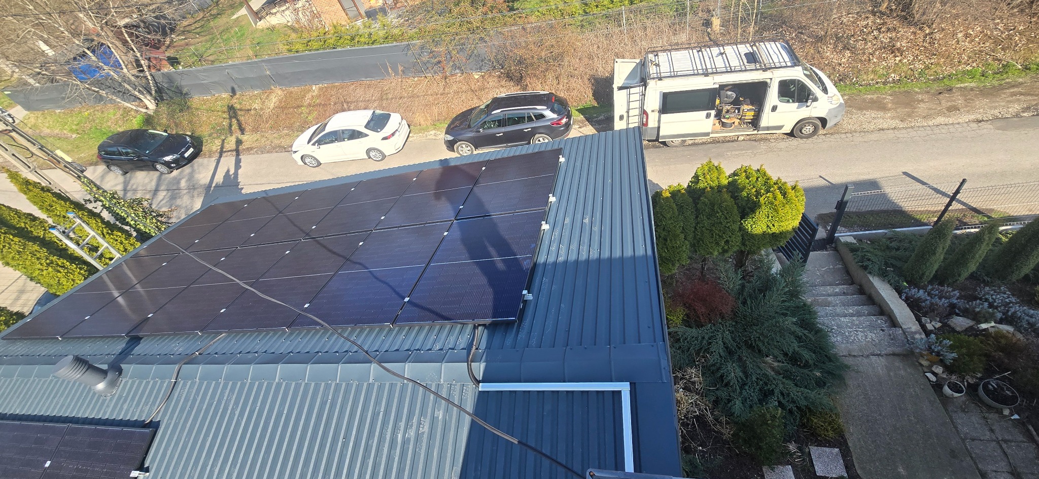 Panel słoneczny na szarym dachu z blachy trapezowej, widok z góry. Na ulicy zaparkowane samochody i bus instalacyjny.