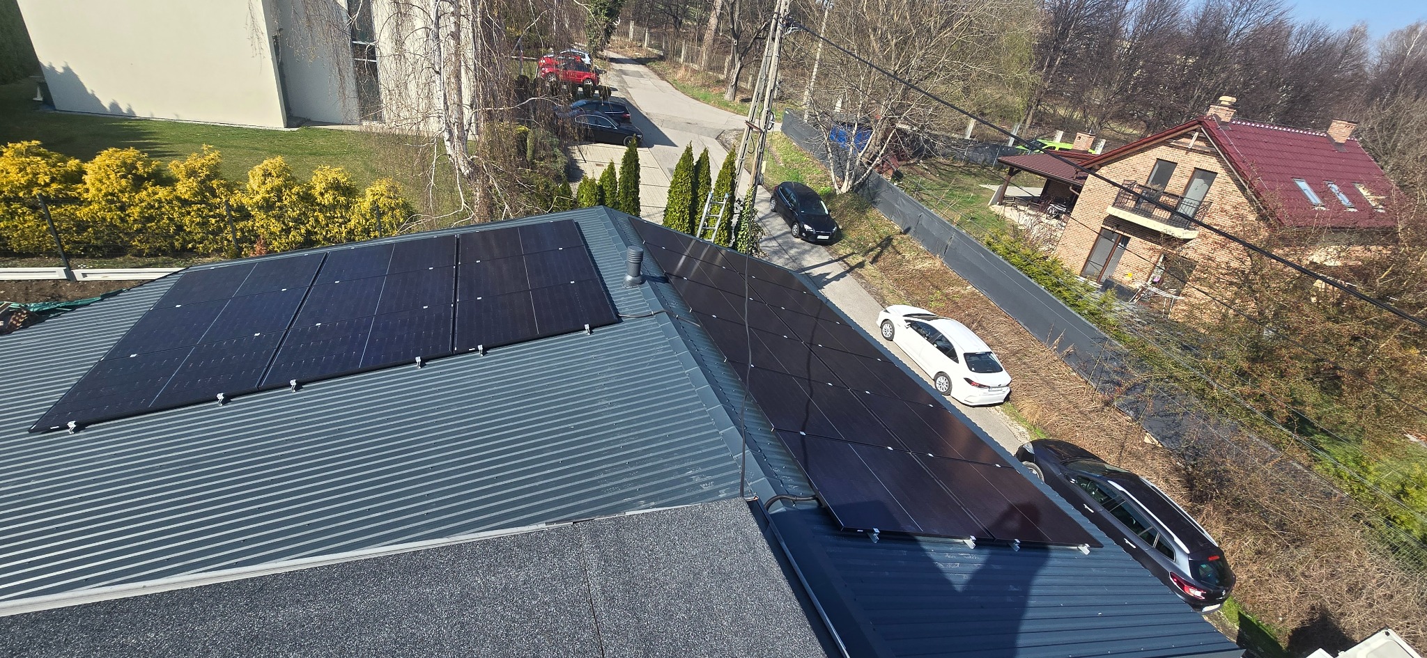 Panele słoneczne na metalowym dachu domu jednorodzinnego, widok z góry, w tle droga z zaparkowanymi samochodami i zabudowa jednorodzinna.