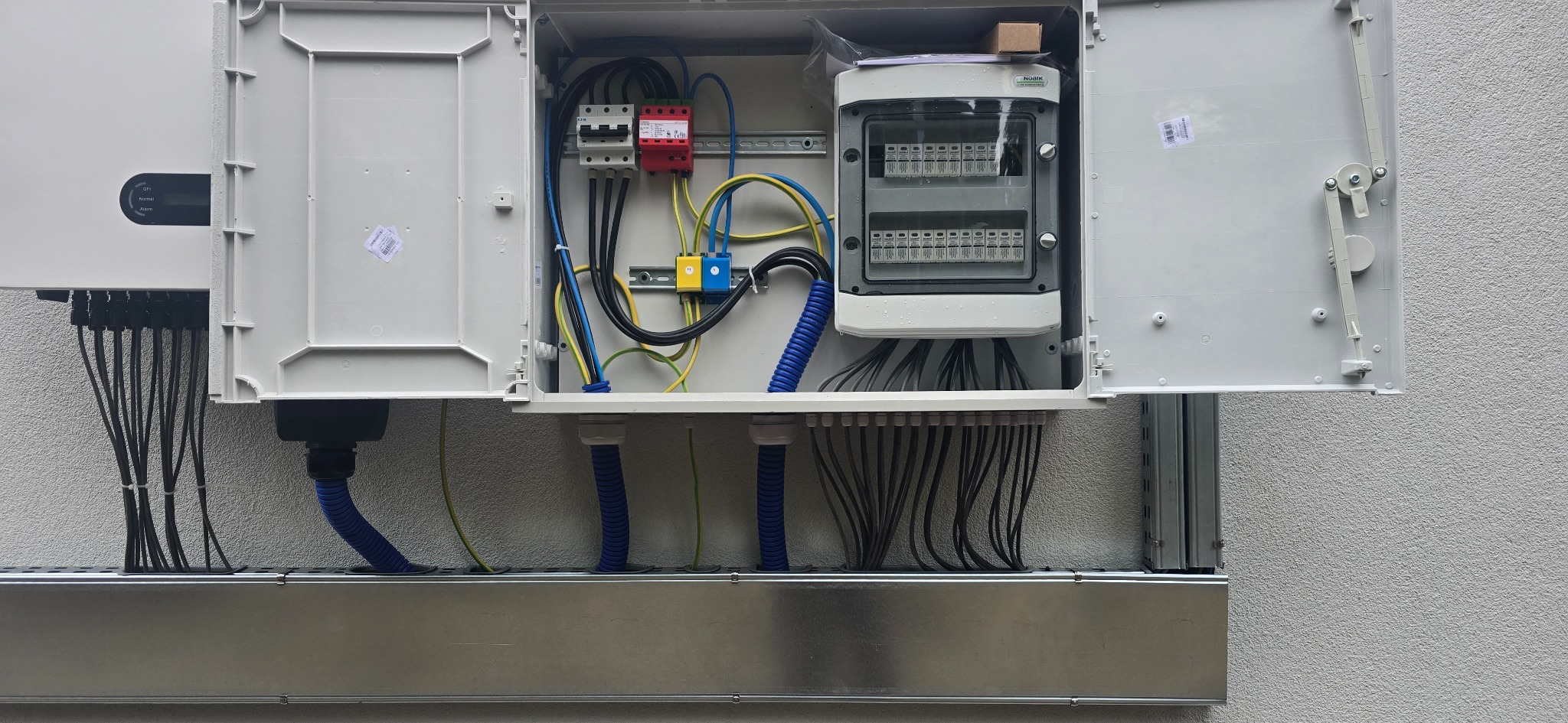 Rozdzielnica z bezpiecznikami, kable w peszlach i listwa montażowa, instalacja fotowoltaiczna na zewnątrz budynku.