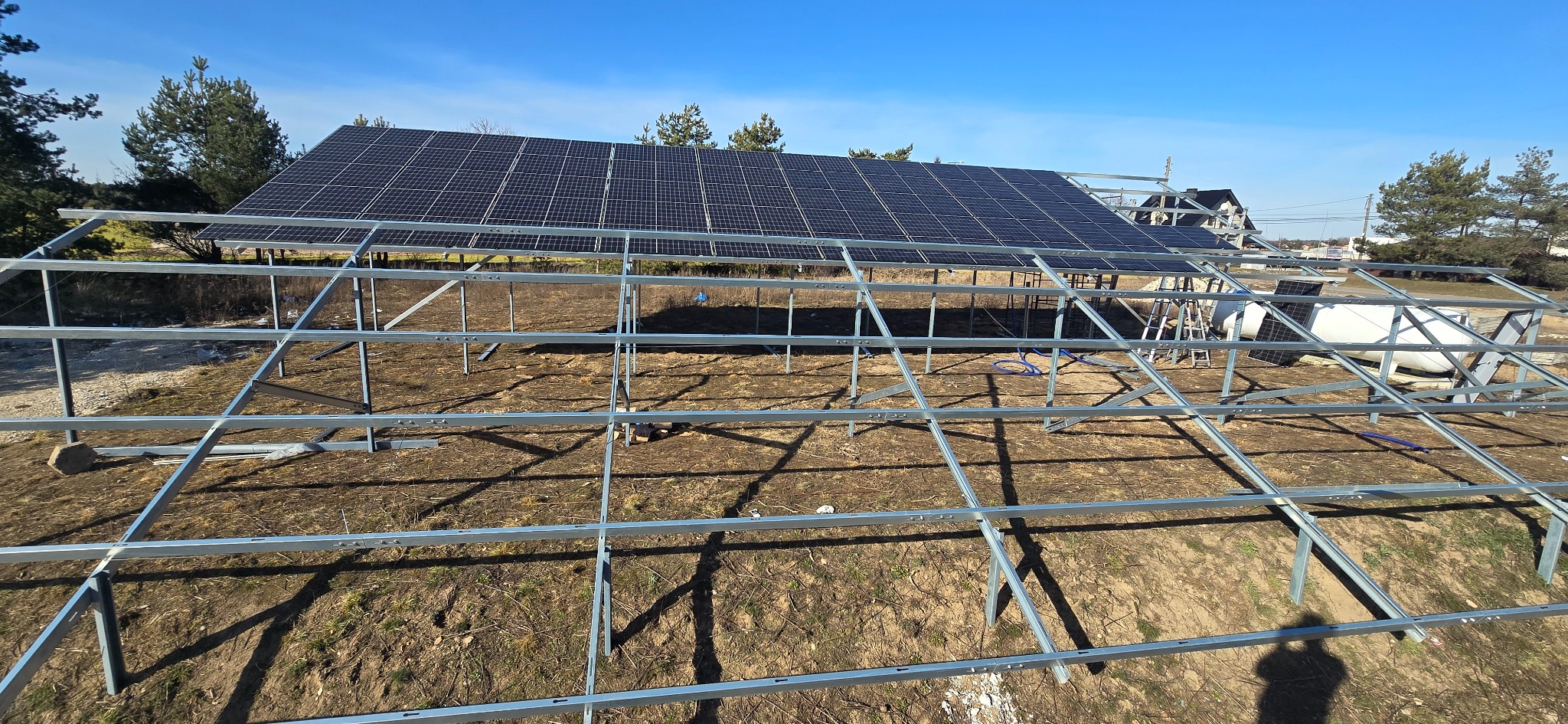 Montaż paneli słonecznych na metalowej konstrukcji. Widok z góry na częściowo zainstalowaną instalację na tle błękitnego nieba i drzew.