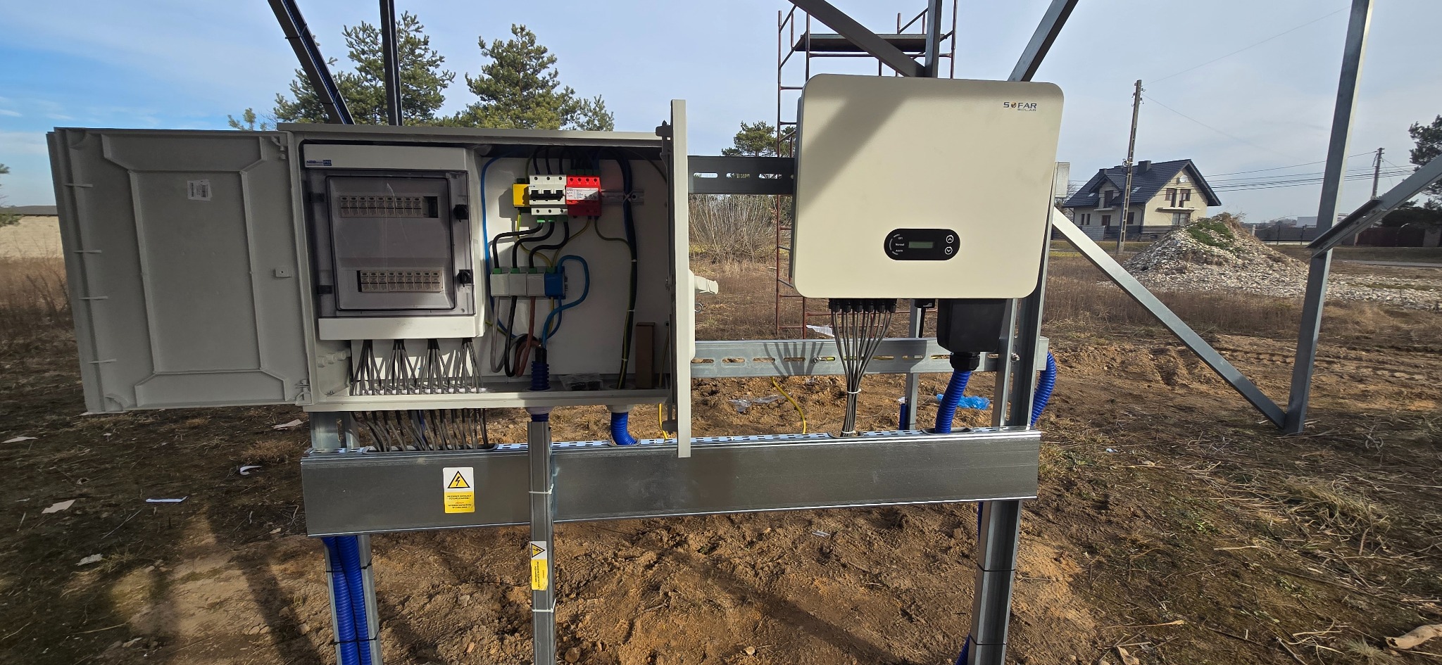 Otwarta skrzynka elektryczna z podłączeniami i falownik Sofar na metalowej konstrukcji, instalacja na zewnątrz budynku. Widoczne kable i zabezpieczenia.
