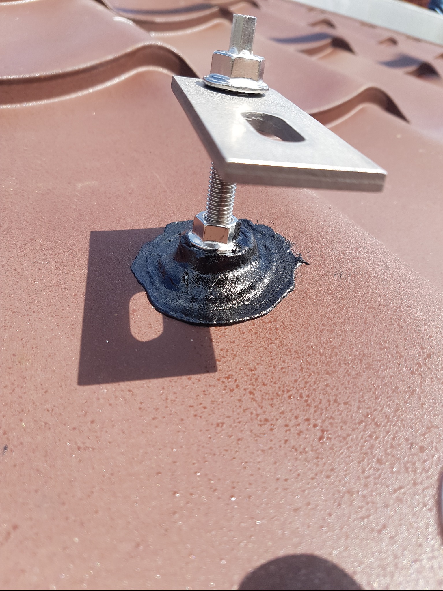 Mocowanie panelu słonecznego na brązowym dachu z dachówki. Uszczelnienie czarnym silikonem. Widok z bliska na pojedynczy uchwyt.