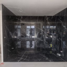 Tomasz Dąbrowski Arsen Usługi Glazurnicze - Czarne płytki imitujące marmur z białymi żyłami, ułożone na ścianach i podłodze w trakcie prac wykończeniowych łazienki. Widoczny profil montażowy i odbicie osoby w płytkach.