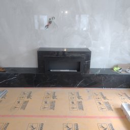 Tomasz Dąbrowski Arsen Usługi Glazurnicze - Montaż czarnego marmurowego kominka elektrycznego na tle białych płytek imitujących marmur. Podłoga zabezpieczona matą budowlaną.