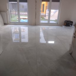 Tomasz Dąbrowski Arsen Usługi Glazurnicze - Błyszcząca, jasna podłoga z płytek imitujących marmur, świeżo położona w pokoju z dużymi oknami. Widoczne detale wykończeniowe i generator prądu w rogu.