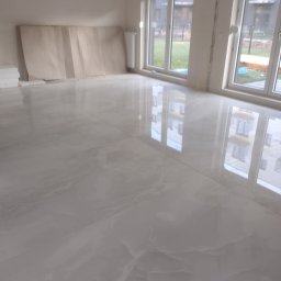 Tomasz Dąbrowski Arsen Usługi Glazurnicze - Połyskująca, jasnoszara podłoga z płytek imitujących marmur w nowym wnętrzu. Widoczne okna z odbiciem budynku, zabezpieczone ściany i stos płytek w rogu.