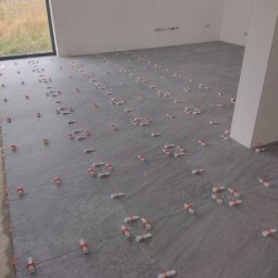 Tomasz Dąbrowski Arsen Usługi Glazurnicze - Szare płytki podłogowe, świeżo ułożone, z widocznymi krzyżykami dystansowymi z czerwonymi paskami, w jasnym, pustym pomieszczeniu z dużym oknem.