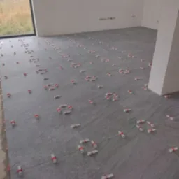 Szare płytki podłogowe, świeżo ułożone, z widocznymi krzyżykami dystansowymi z czerwonymi paskami, w jasnym, pustym pomieszczeniu z dużym oknem.