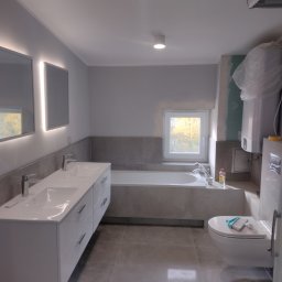 Tomasz Dąbrowski Arsen Usługi Glazurnicze - Nowoczesna łazienka w szarościach: podwójna umywalka z szafką, wanna w zabudowie, toaleta wisząca. Widoczne płytki podłogowe i ścienne, okno. Część prac wykończeniowych w toku.