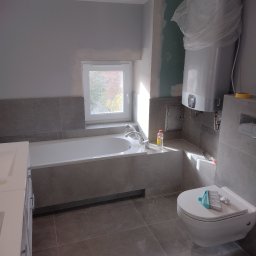 Tomasz Dąbrowski Arsen Usługi Glazurnicze - Szara łazienka w trakcie remontu: wanna zabudowana płytkami, podwieszana toaleta z szczotką, bojler w folii. Widoczne niedokończone ściany i okno.