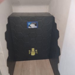 Tomasz Dąbrowski Arsen Usługi Glazurnicze - Częściowo wykończona łazienka z czarnymi, heksagonalnymi płytkami na ścianie i beżowymi płytkami imitującymi drewno na podłodze. Widoczna instalacja podtynkowa WC.