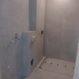 Szara łazienka w trakcie wykańczania: ułożone płytki podłogowe z systemem poziomowania, widoczne instalacje elektryczne i wodne, surowy stan ścian.