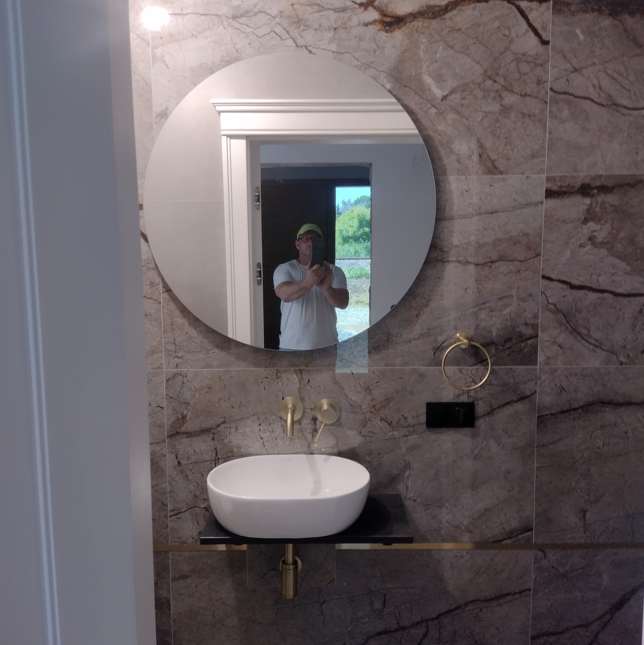 Wykończona łazienka z umywalką nablatową, armaturą w kolorze złota i dużym okrągłym lustrem, w którym odbija się osoba robiąca zdjęcie.