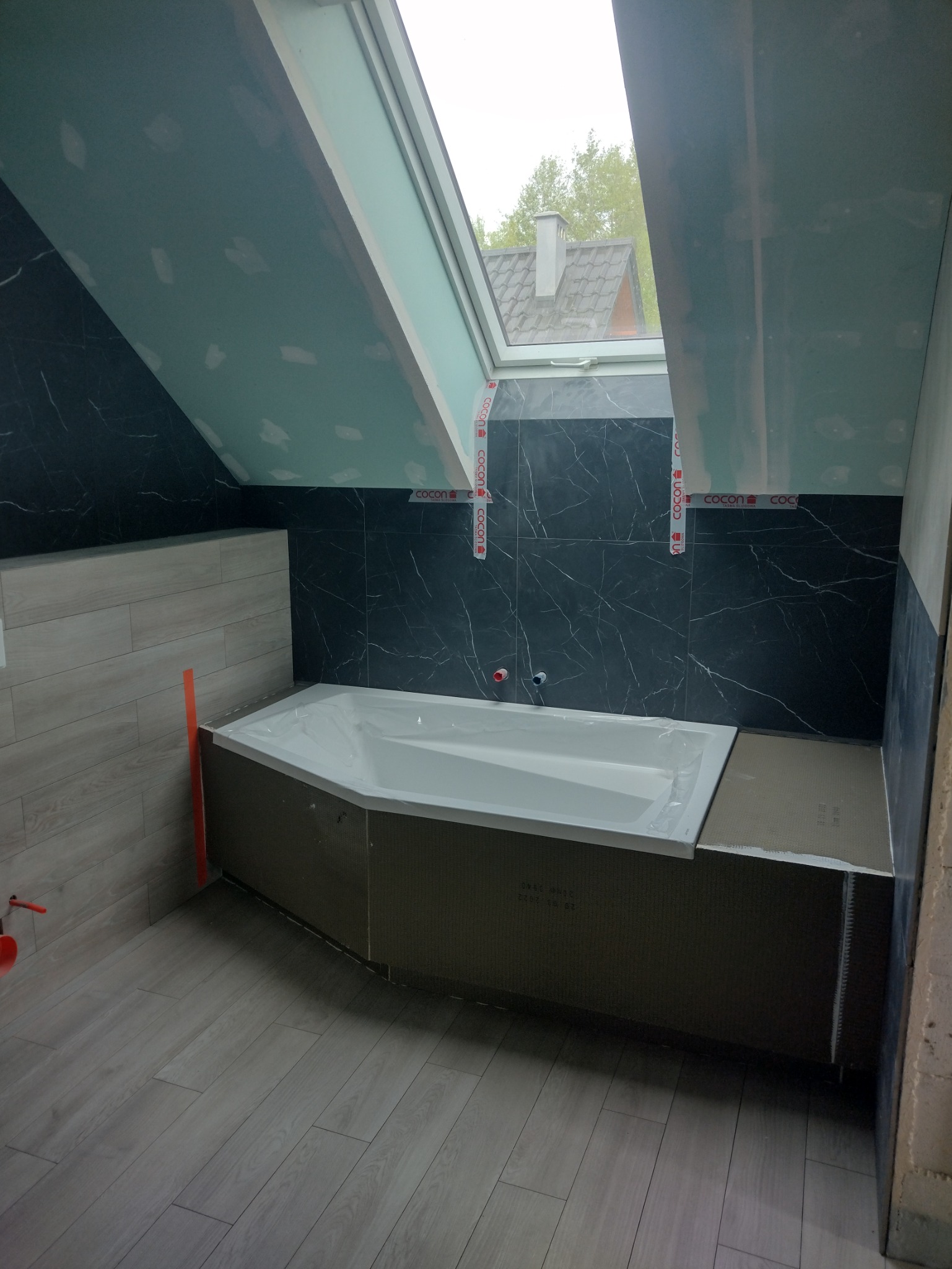 Łazienka na poddaszu z oknem dachowym w trakcie wykańczania. Widoczna wanna obudowana płytami, położona glazura imitująca drewno i marmur.