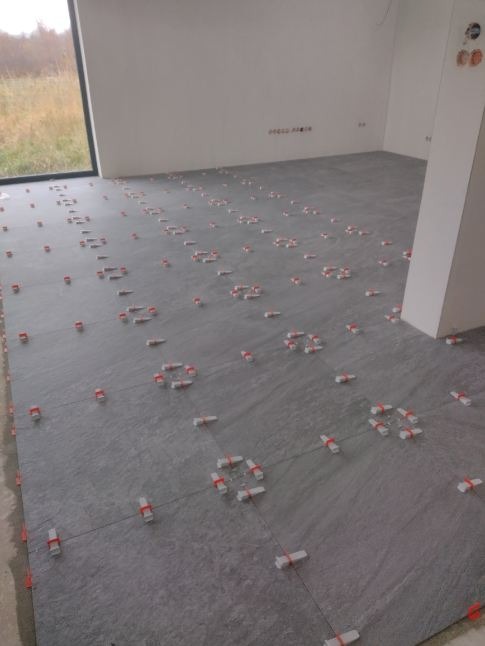 Szare płytki podłogowe, świeżo ułożone, z widocznymi krzyżykami dystansowymi z czerwonymi paskami, w jasnym, pustym pomieszczeniu z dużym oknem.