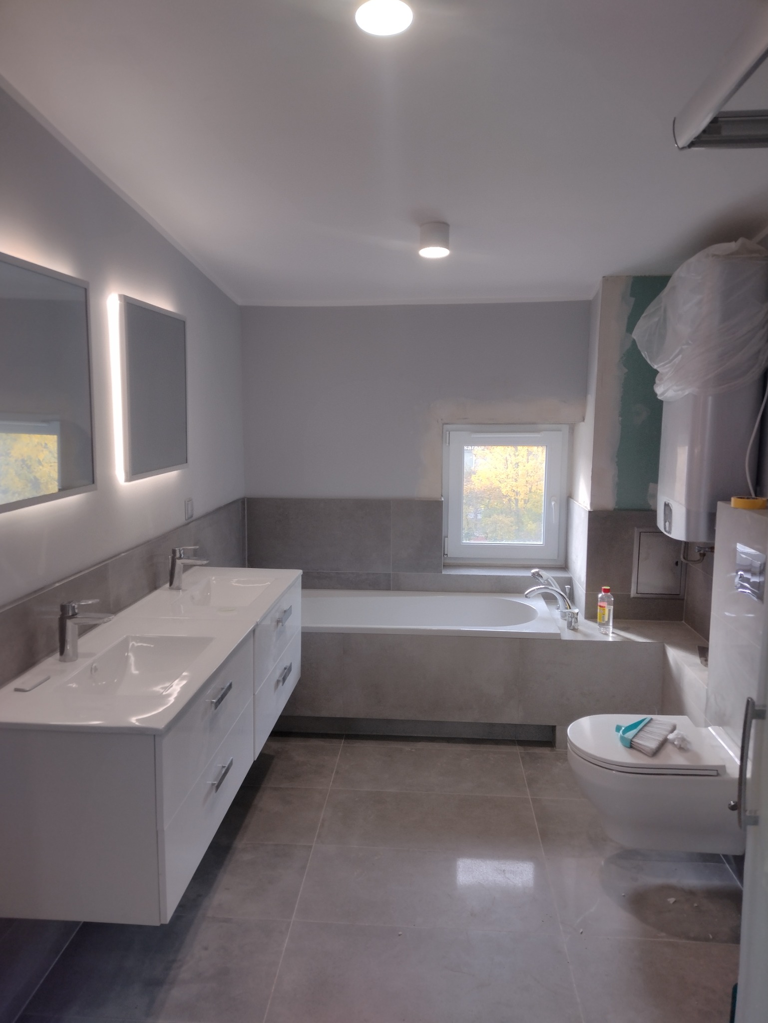 Nowoczesna łazienka w szarościach: podwójna umywalka z szafką, wanna w zabudowie, toaleta wisząca. Widoczne płytki podłogowe i ścienne, okno. Część prac wykończeniowych w toku.