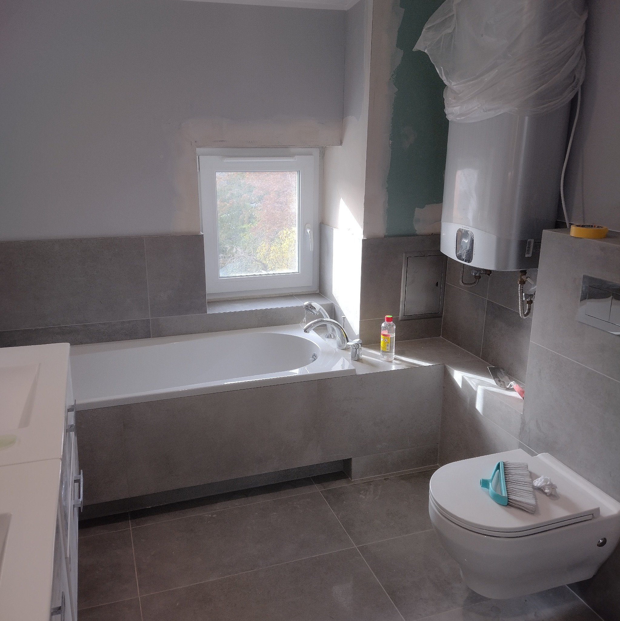 Szara łazienka w trakcie remontu: wanna zabudowana płytkami, podwieszana toaleta z szczotką, bojler w folii. Widoczne niedokończone ściany i okno.