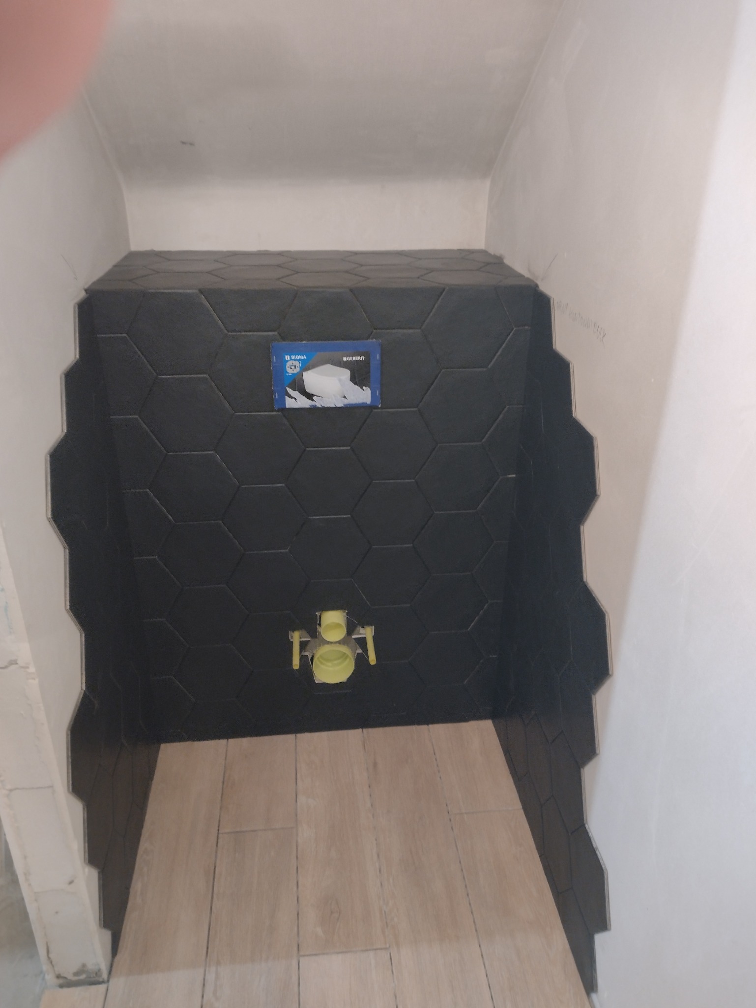 Częściowo wykończona łazienka z czarnymi, heksagonalnymi płytkami na ścianie i beżowymi płytkami imitującymi drewno na podłodze. Widoczna instalacja podtynkowa WC.