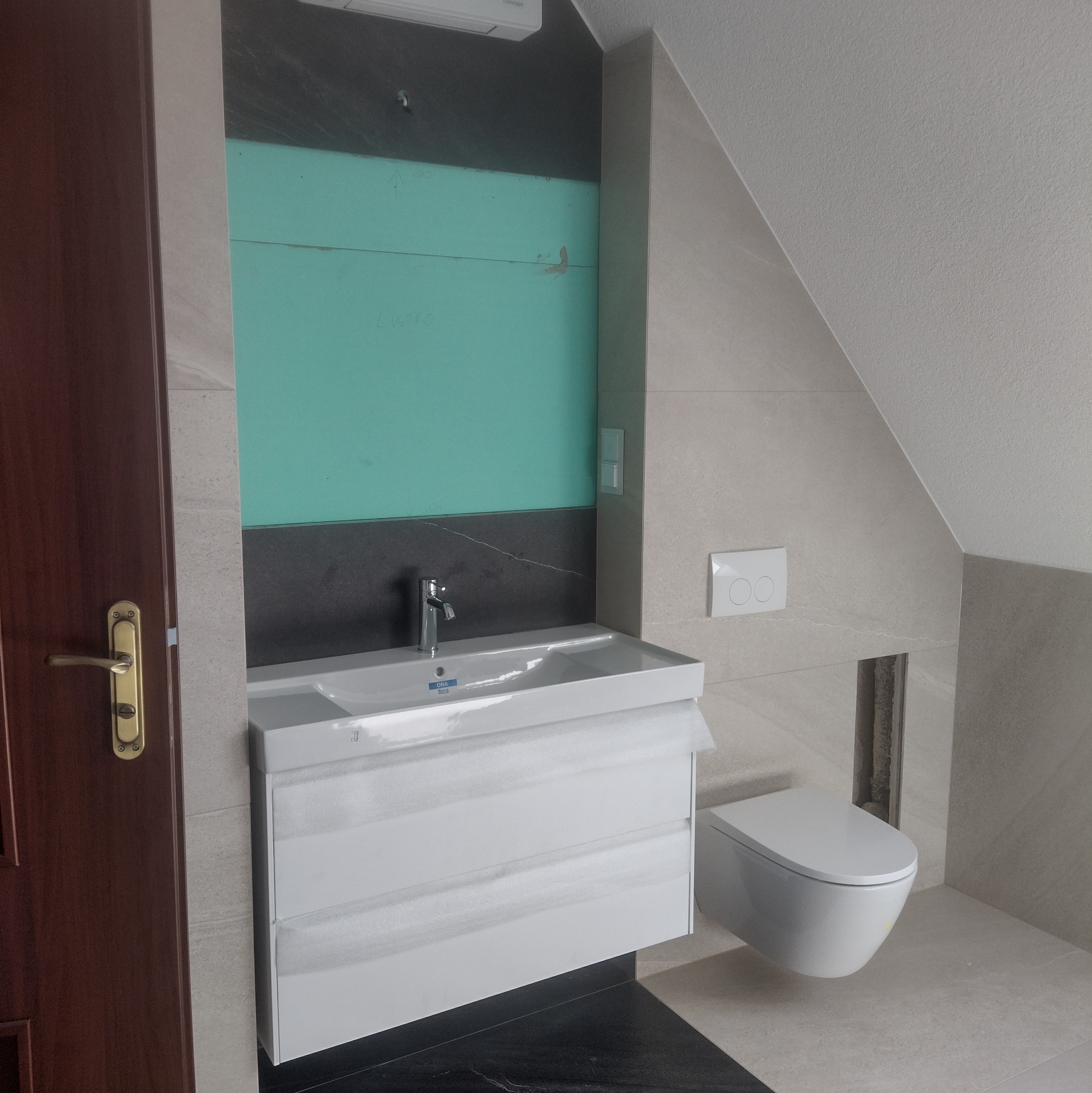 Nowoczesna łazienka w trakcie wykańczania: podwieszana toaleta i szafka z umywalką. Część ściany z płytami GK i widoczną instalacją. Ciemne płytki podłogowe.