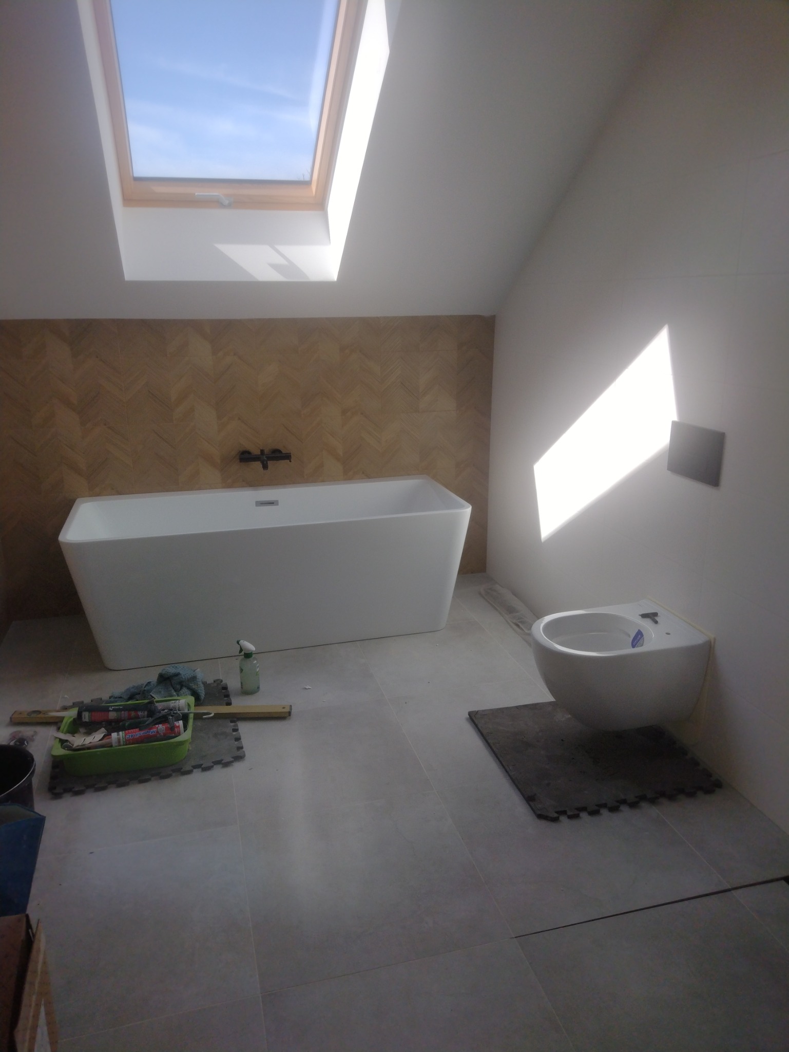 Nowoczesna łazienka na poddaszu z oknem dachowym: biała wanna na tle ściany w jodełkę, wisząca toaleta. Na podłodze szare płytki i narzędzia glazurnicze.