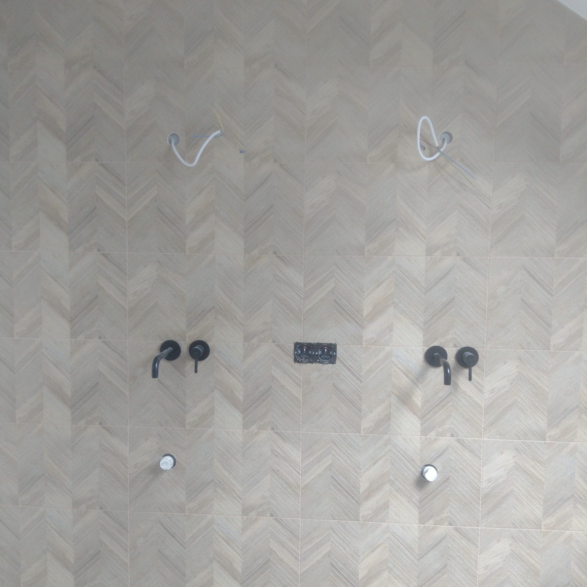 Ściana łazienki wyłożona jasnymi płytkami w jodełkę. Widoczne wystające kable, czarne baterie i elementy instalacji podtynkowej. Etap prac wykończeniowych.