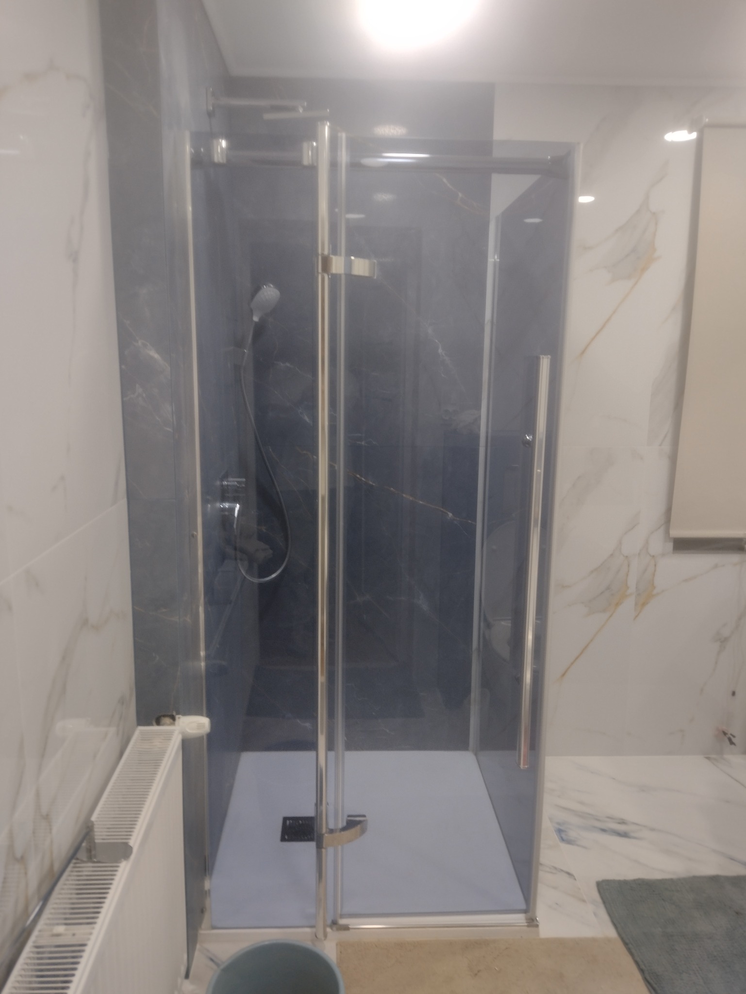 Nowoczesna kabina prysznicowa z szarymi płytkami imitującymi marmur i chromowanymi profilami, widoczna w łazience z marmurową podłogą.