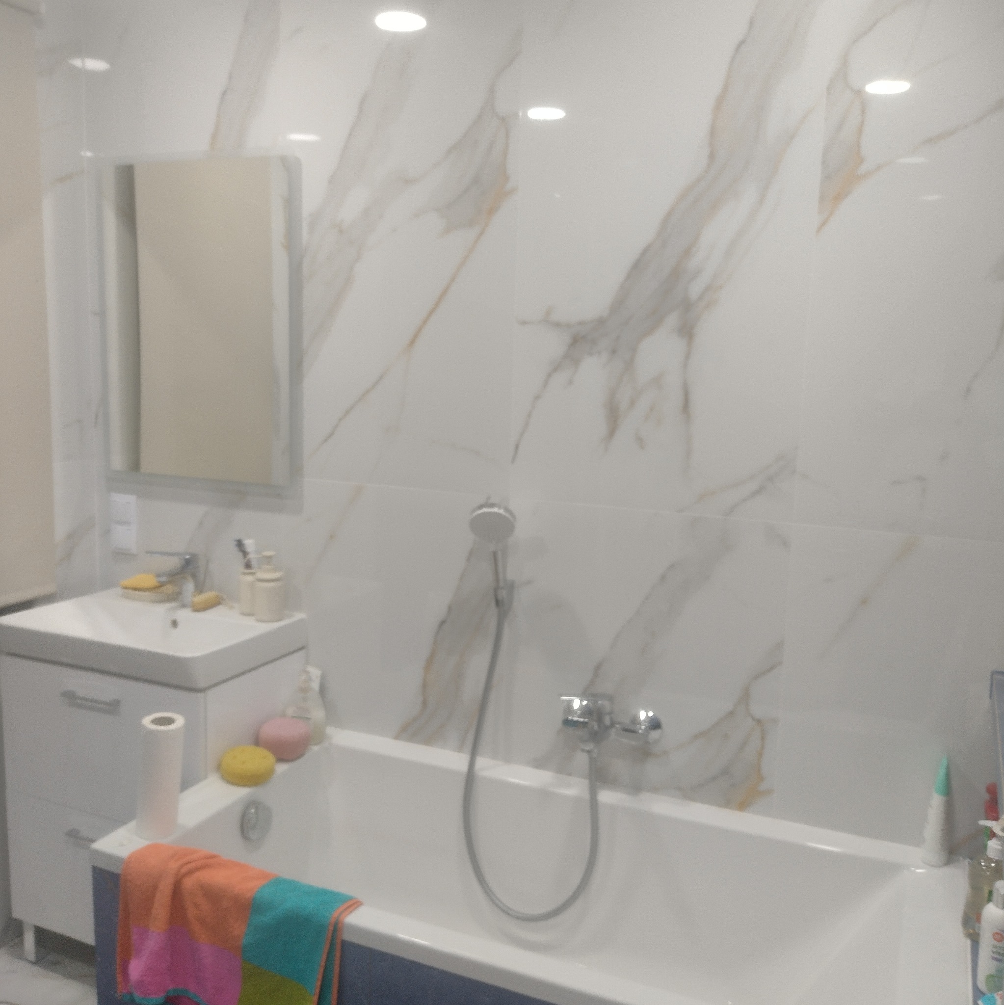 Biała łazienka z wanną, umywalką i lustrem. Ściany wyłożone płytkami imitującymi marmur z delikatnymi złotymi żyłkami. Na wannie leżą kolorowe ręczniki i akcesoria.