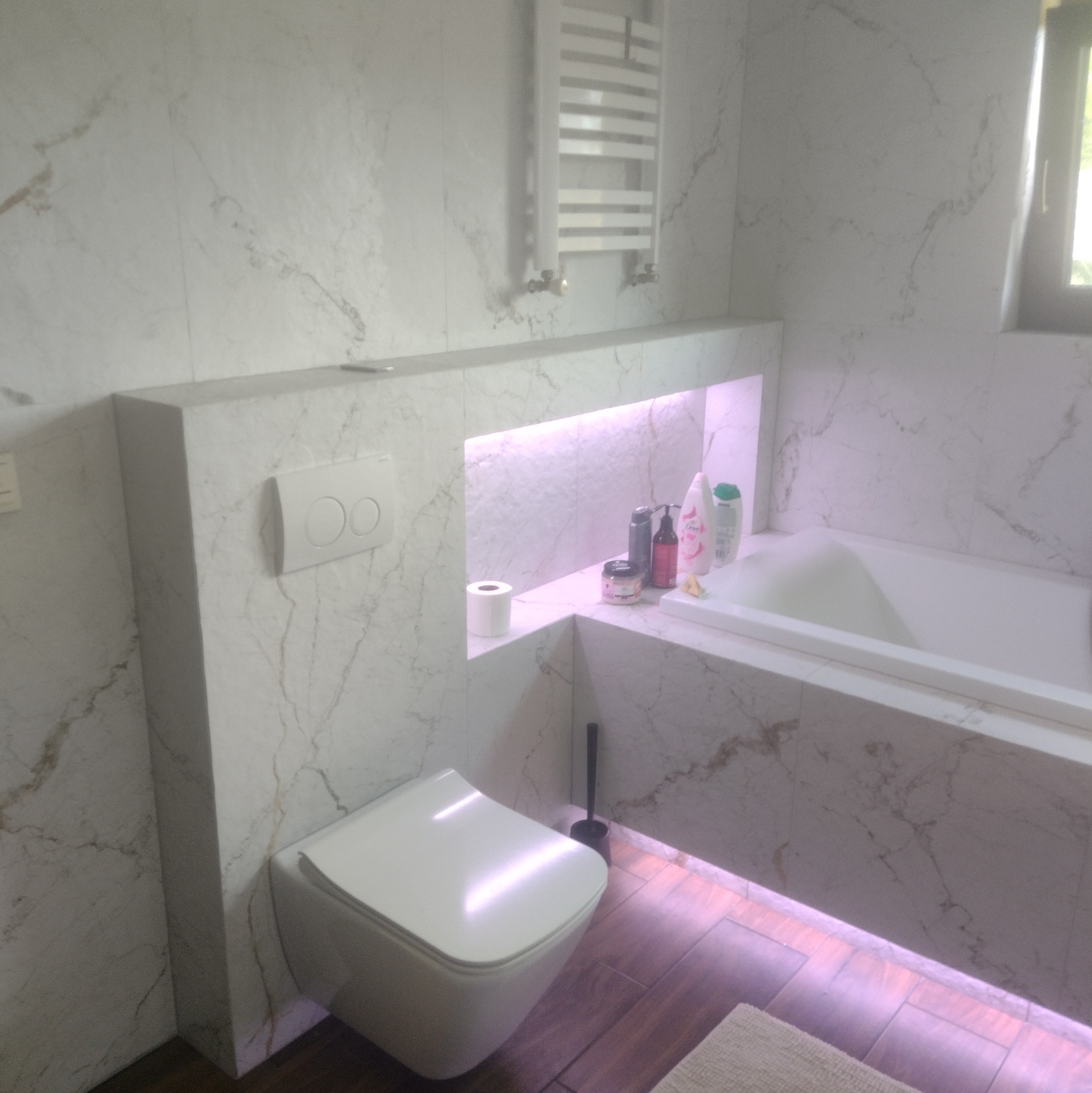 Elegancka łazienka z marmurowymi płytkami, podświetlaną wnęką nad wanną i podwieszaną toaletą. Minimalistyczny grzejnik drabinkowy i drewnopodobna podłoga.