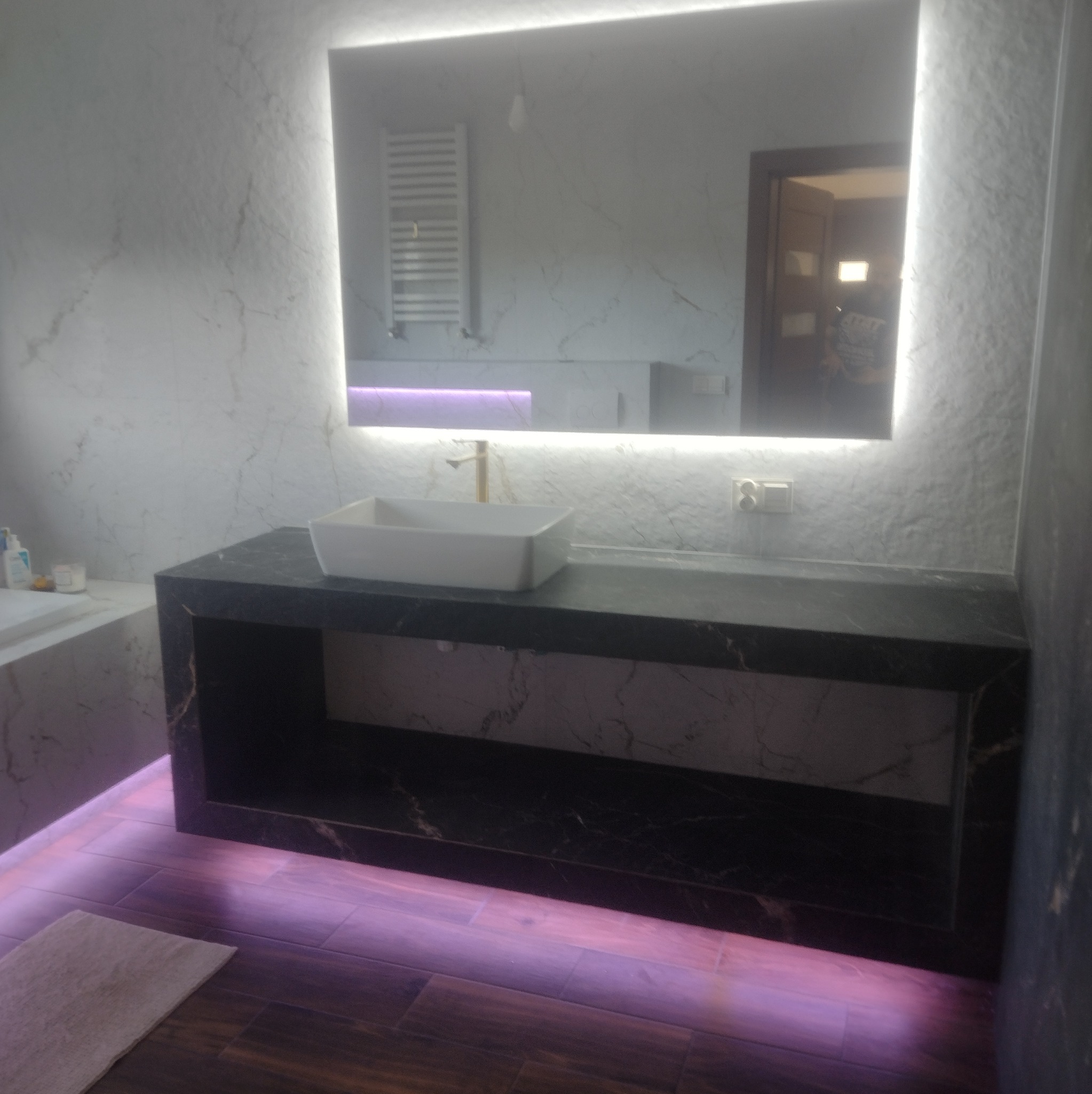 Nowoczesna łazienka z podświetlanym lustrem, marmurową ścianą i czarnym blatem umywalki. Podłoga z drewnopodobnych paneli z fioletowym podświetleniem.