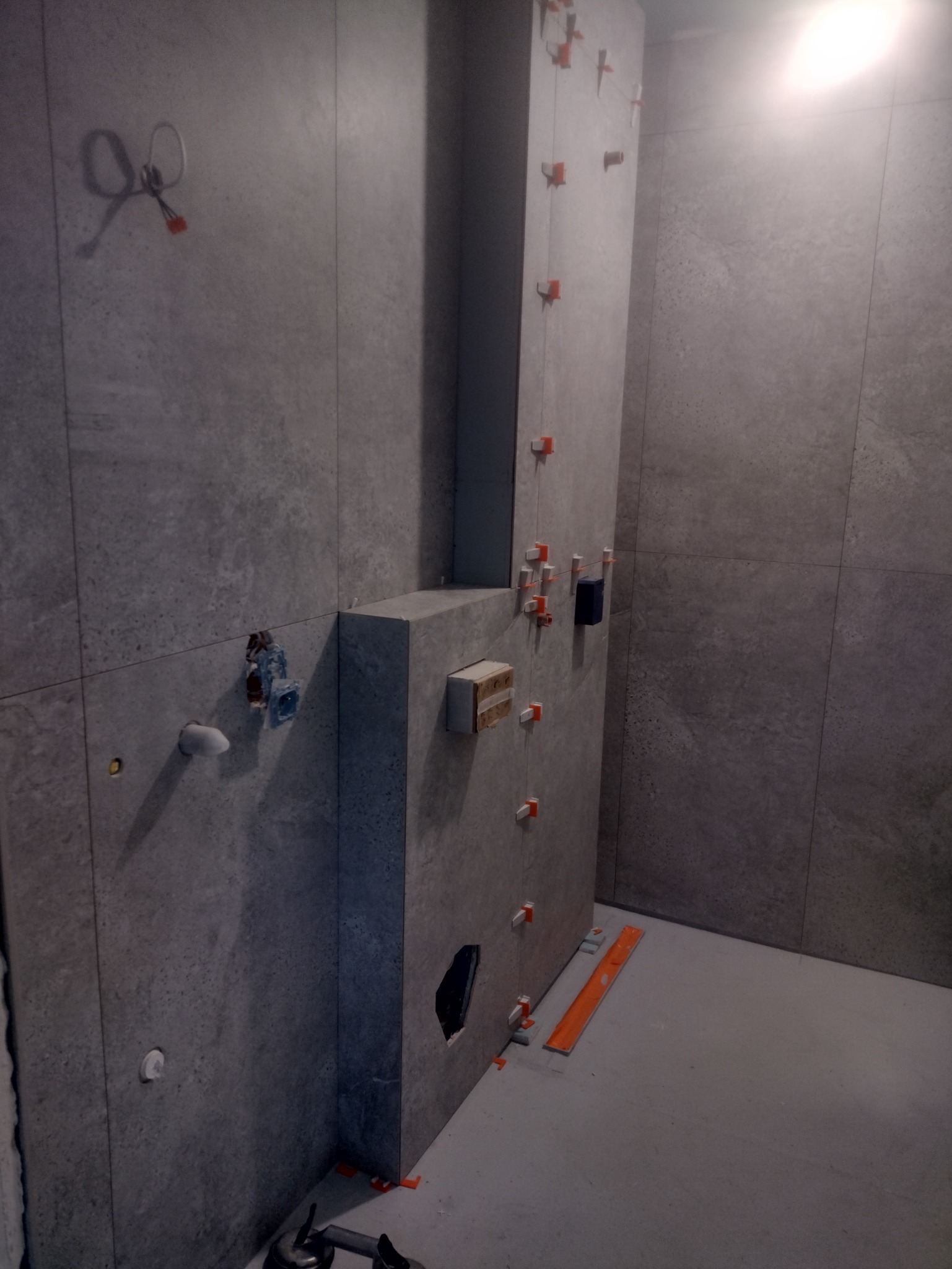 Szara łazienka w trakcie wykończenia: widoczne płytki, poziomica i elementy instalacji. Na ścianach krzyżyki glazurnicze, sugerujące świeżo położone płytki.