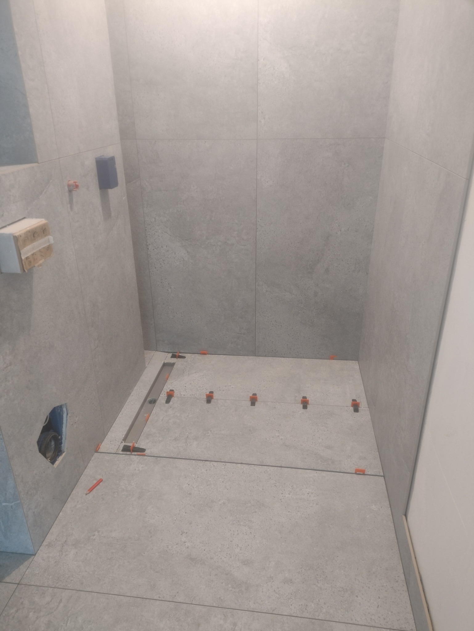 Szare płytki w łazience w trakcie montażu. Widoczny odpływ liniowy, pomarańczowe klipsy poziomujące, fragment puszki elektrycznej i ołówek na podłodze.