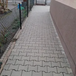 Szara kostka brukowa ułożona wzdłuż ogrodzenia i ściany budynku. Widok z góry, perspektywa zbieżna, oświetlenie dzienne.