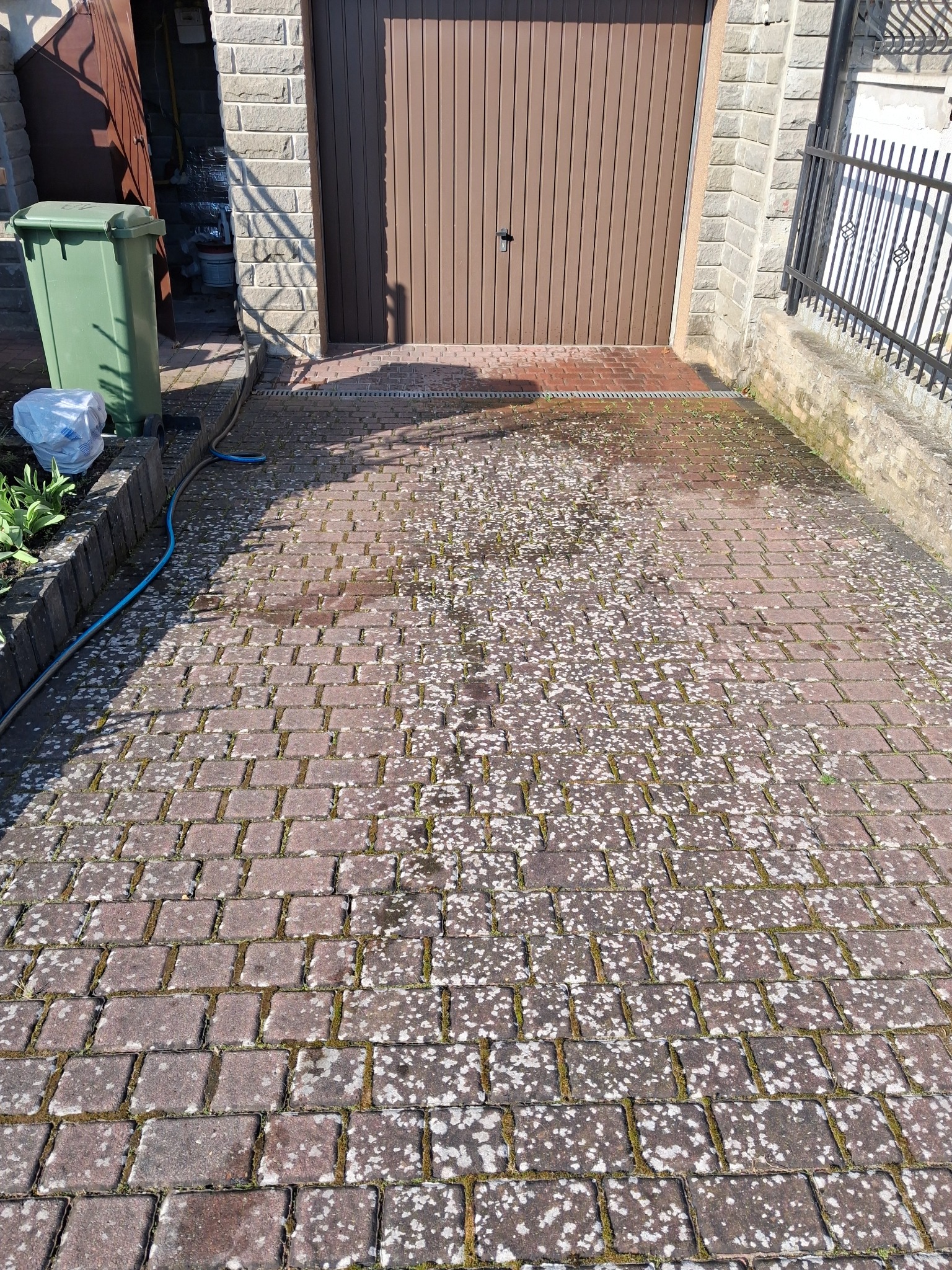 Częściowo umyta, czerwona kostka brukowa przed garażem, z widocznymi śladami białego nalotu i mchu, z niebieskim wężem ogrodowym w pobliżu ścieku.