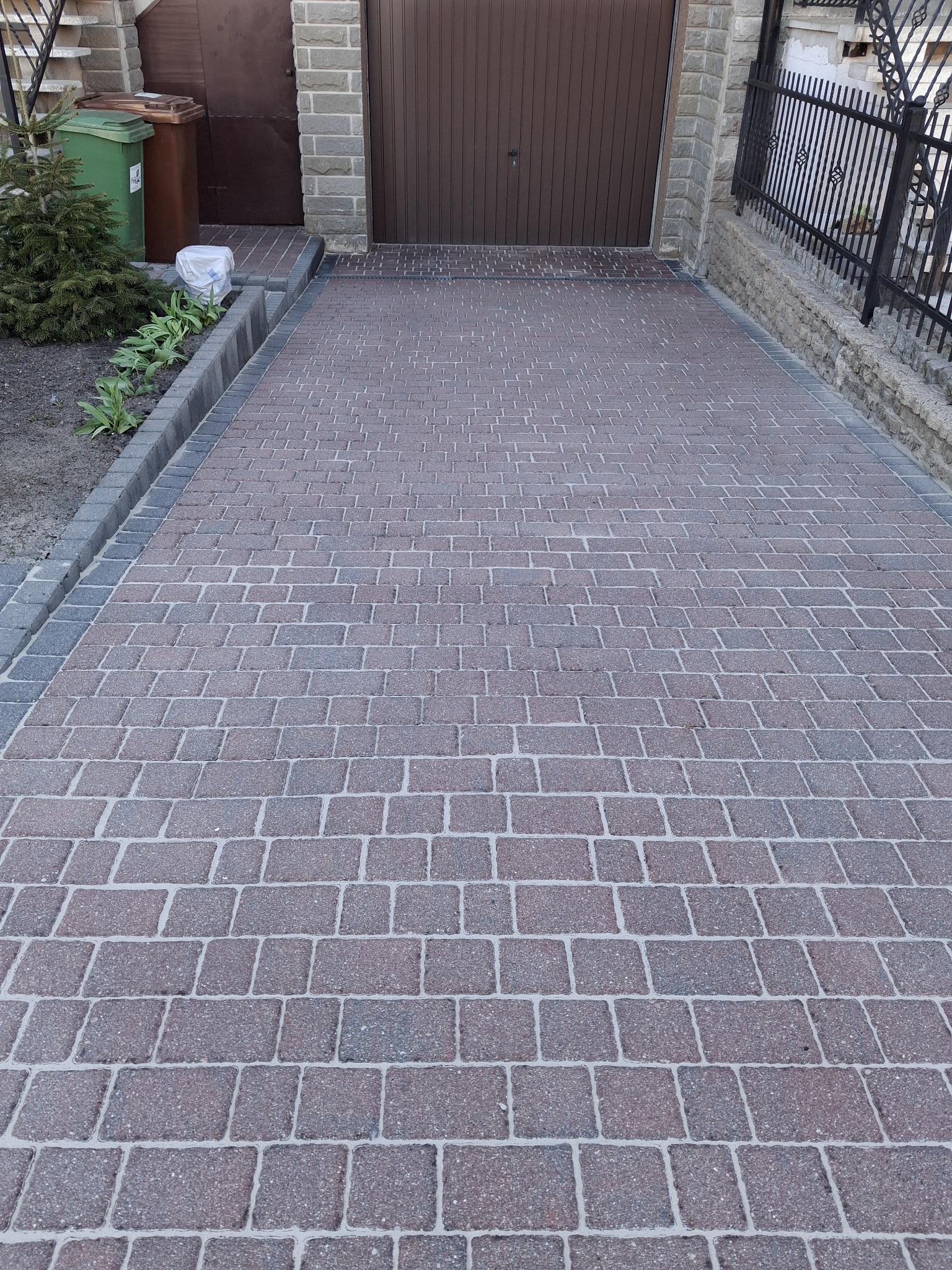 Czerwono-szara kostka brukowa ułożona na podjeździe przed garażem, z widocznymi obrzeżami i elementami zieleni po bokach. Widok z góry na regularny wzór kostki.