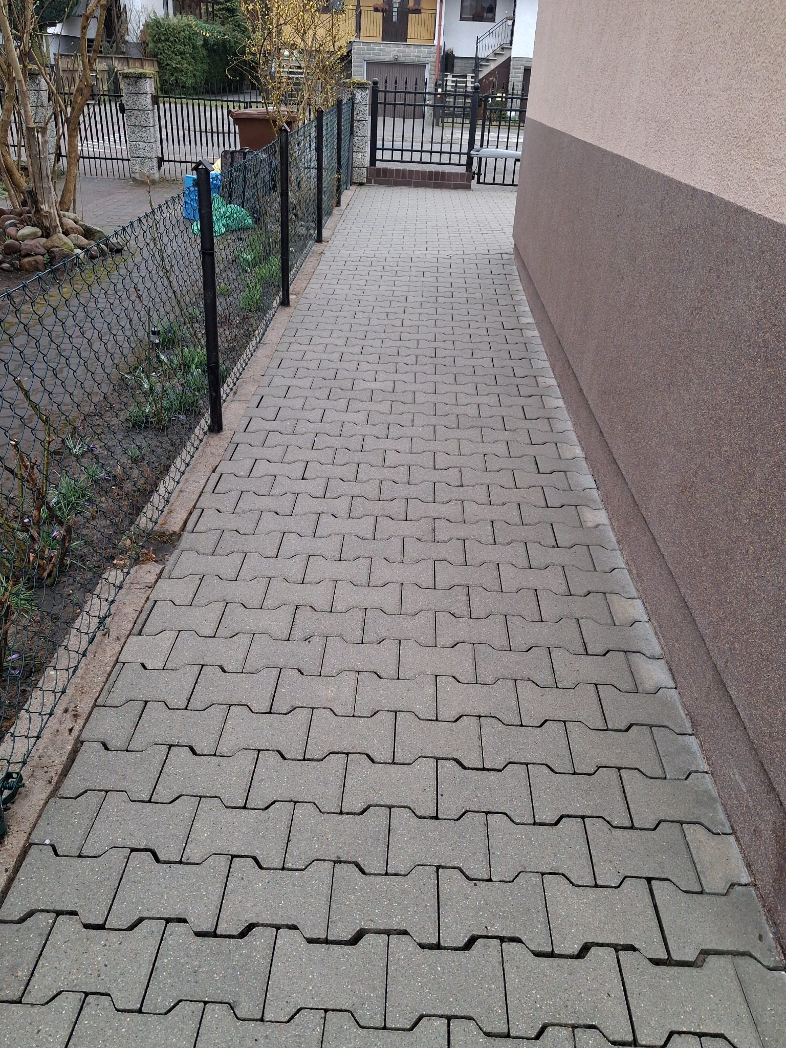 Szara kostka brukowa ułożona wzdłuż ogrodzenia i ściany budynku. Widok z góry, perspektywa zbieżna, oświetlenie dzienne.