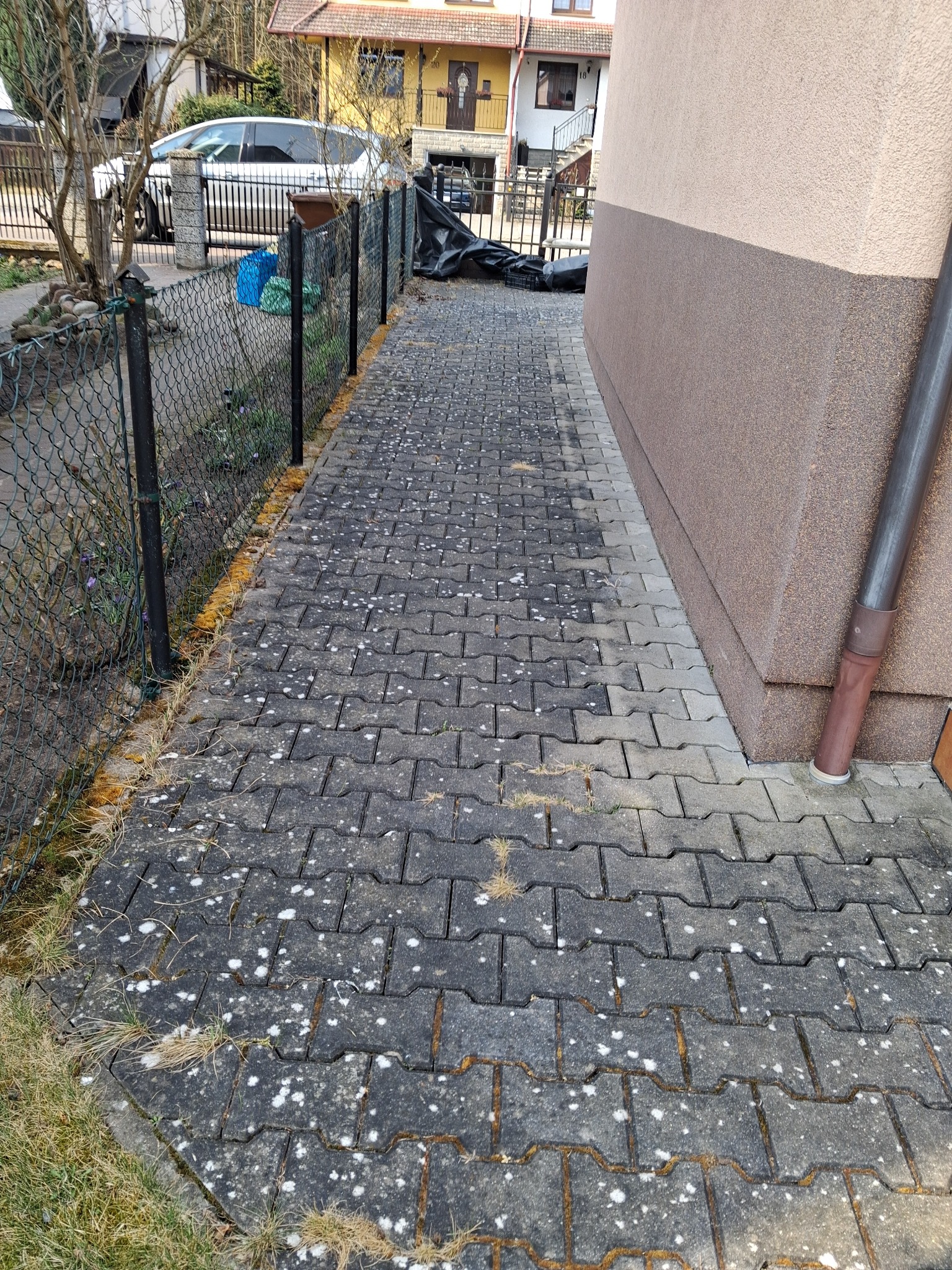 Brudna kostka brukowa przed budynkiem, widoczne zabrudzenia biologiczne i mech. Część kostki jest zacieniona, a część oświetlona. Obok ogrodzenie i ściana budynku.