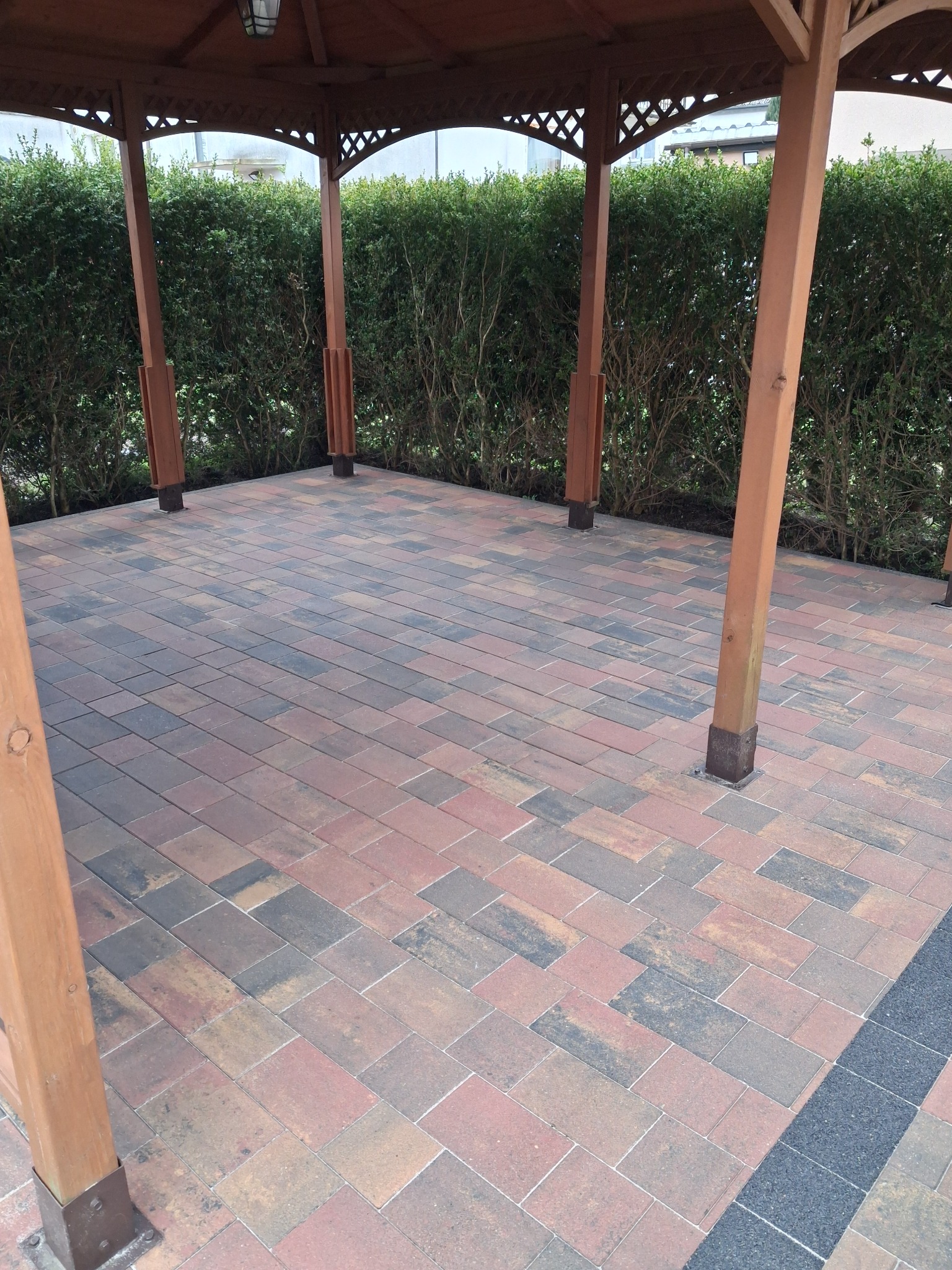 Kolorowa kostka brukowa pod drewnianą altaną, otoczona zielenią. Widoczne detale łączeń i różnorodność odcieni kostki. Estetyczne wykończenie.