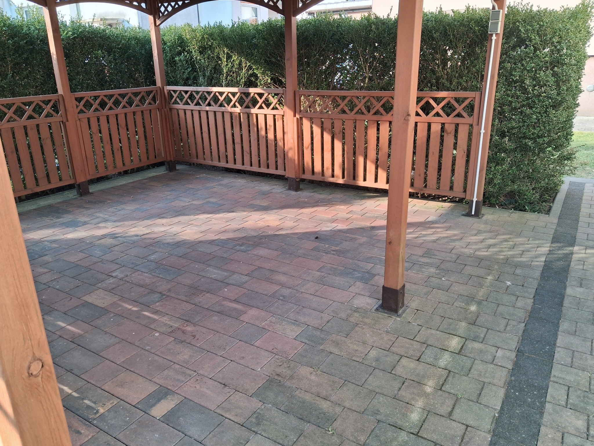 Częściowy widok zadaszonej altany z drewnianą konstrukcją i brukowaną podłogą. Widoczne zielone krzewy w tle i fragment chodnika z kostki brukowej.