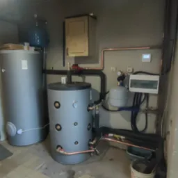 Instalacja dwóch szarych zbiorników ciepłej wody z miedzianymi rurami w kotłowni z uszkodzoną podłogą z płytek i widocznymi elementami sterowania.