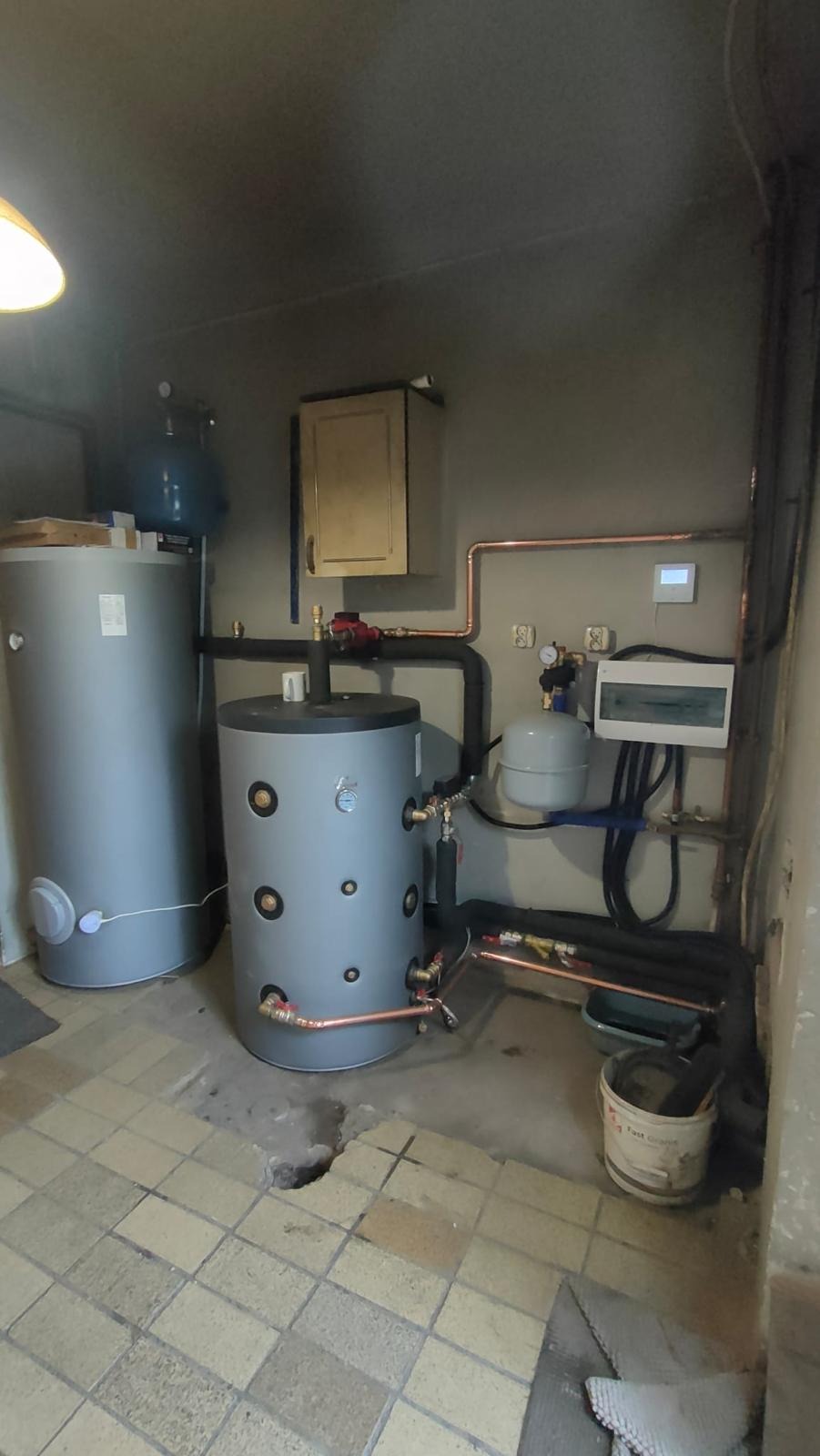 Instalacja dwóch szarych zbiorników ciepłej wody z miedzianymi rurami w kotłowni z uszkodzoną podłogą z płytek i widocznymi elementami sterowania.