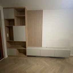 Almal - Jasna zabudowa salonu z drewnianymi lamelami i półkami, podłużna szafka RTV, parkiet w jodełkę. Minimalistyczny design, ciepłe oświetlenie.