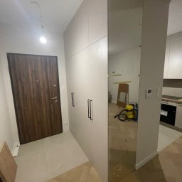 Almal - Jasna szafa w przedpokoju z lustrzanym frontem, odbijająca wnętrze mieszkania. Widoczna drewniana podłoga i drzwi wejściowe. Minimalistyczny design.