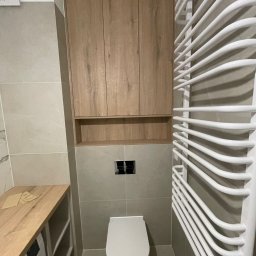 Almal - Drewniana szafka łazienkowa nad toaletą, z półką i trzema drzwiczkami, obok grzejnik drabinkowy. Umywalka z szafką w tle. Szare płytki na ścianach i podłodze.