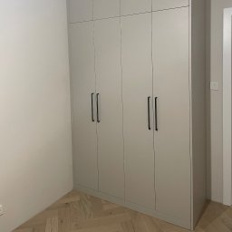 Almal - Jasnoszara, czterodrzwiowa szafa wnękowa z podłużnymi, czarnymi uchwytami, na parkiecie w jodełkę, w rogu pokoju z włącznikiem światła na ścianie.