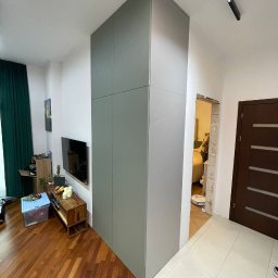 Almal - Szara, wysoka szafa wnękowa od podłogi do sufitu, zlicowana ze ścianą, w nowoczesnym salonie z drewnianą podłogą i telewizorem na ścianie.