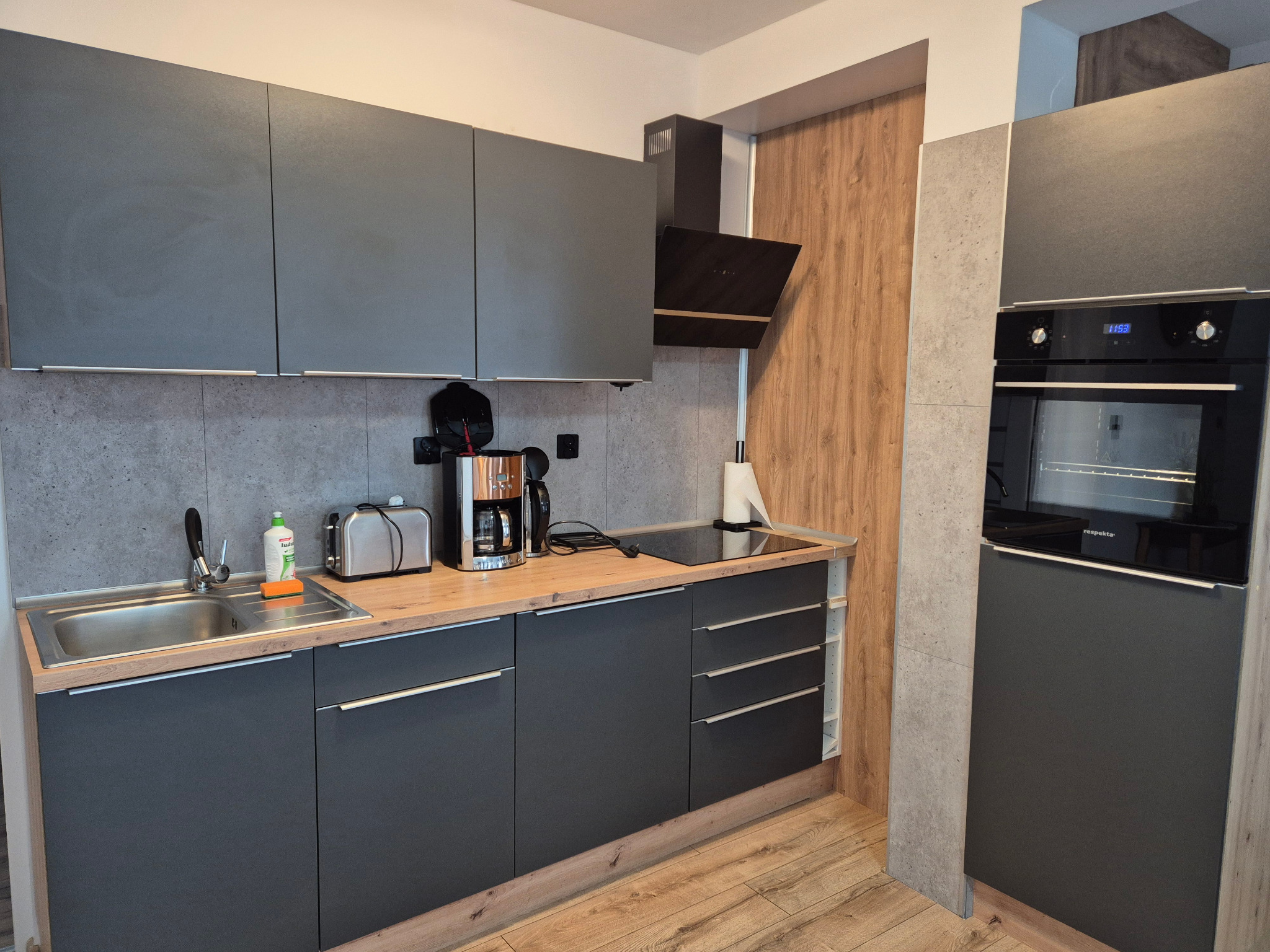 Nowoczesna, szara kuchnia z drewnianymi akcentami. Widoczny zlew z płynem, toster, ekspres i piekarnik. Ujęcie wnętrza po sprzątaniu.